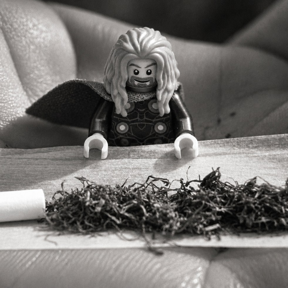 The caped figure stands in an open palm behind a rolling paper spread with tobacco